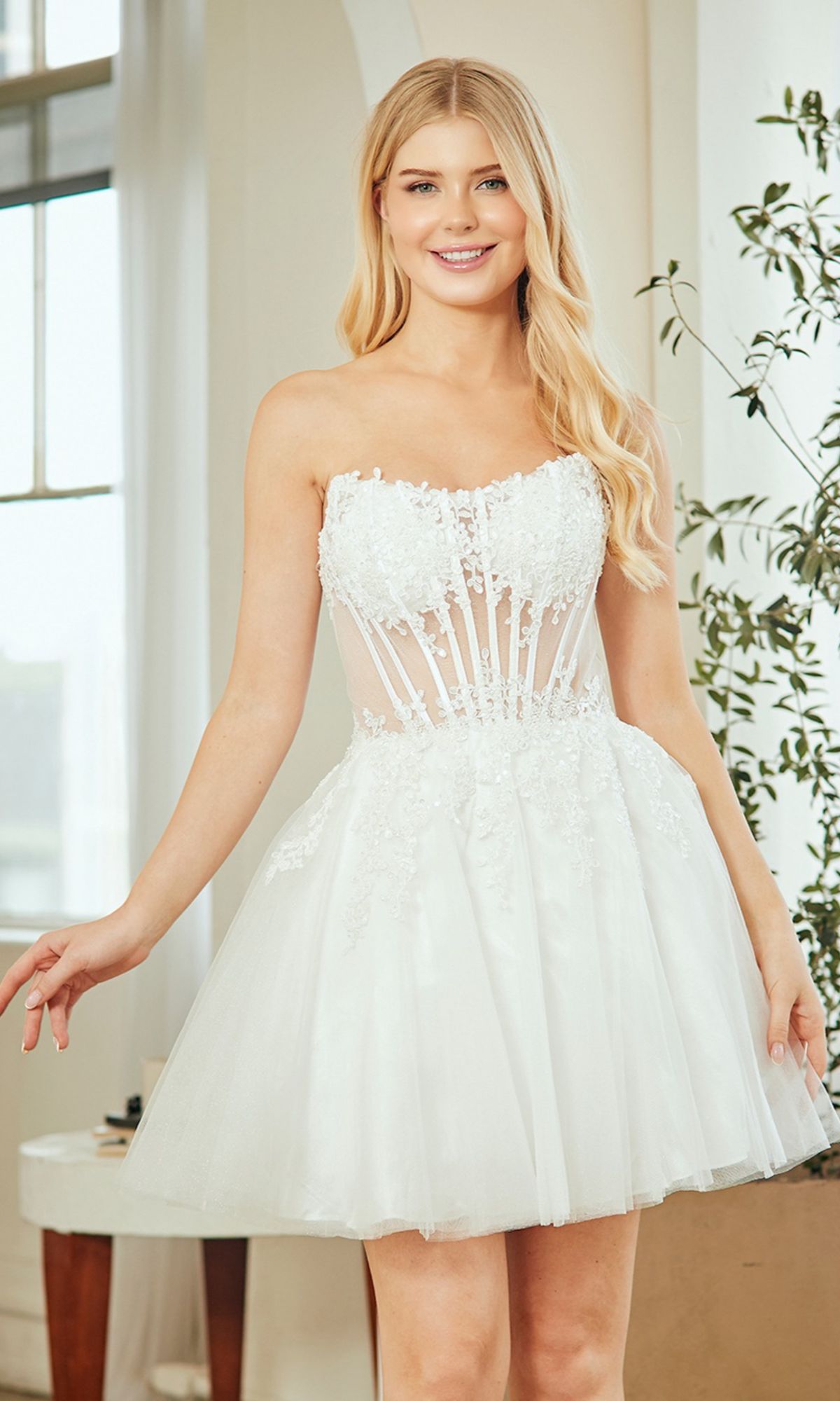 A woman in the Amelia Couture 9083S Strapless Short Party Dress with a sheer, floral-embellished bodice and tulle skirt stands by a window and potted plant—an ideal chic choice for homecoming.